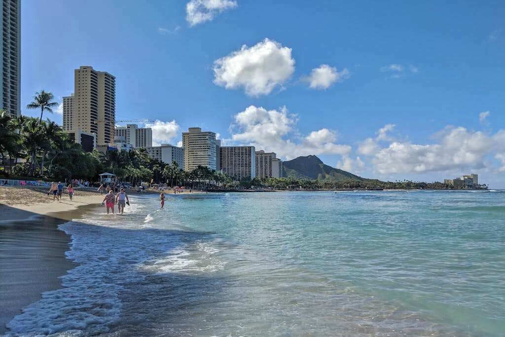 A beach with clear water, palm trees, and high-rise buildings in the background.