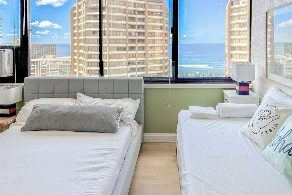 A hotel room with two beds, a large window with a city view, a bedside table with a lamp, and a nightstand with a phone.