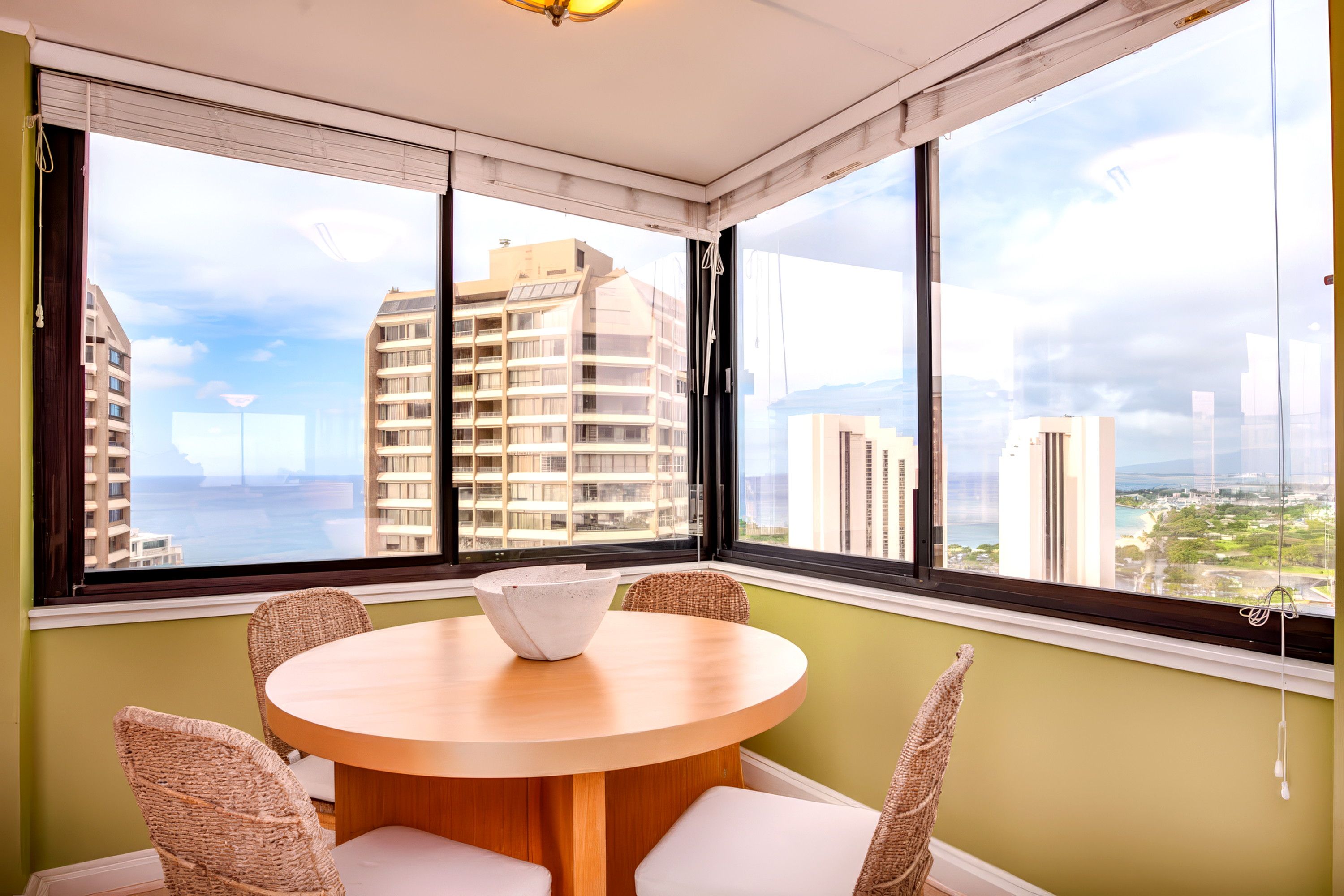 A room with a round table, four wicker chairs, and large windows offering a city view.