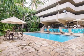 A swimming pool with lounge chairs and umbrellas, surrounded by greenery and a multi-story building with a blue mural. - Windsor Tower Waikiki Apartment | A/C, Laundry, Pool, Xbox | One Block to Beach (Honolulu)