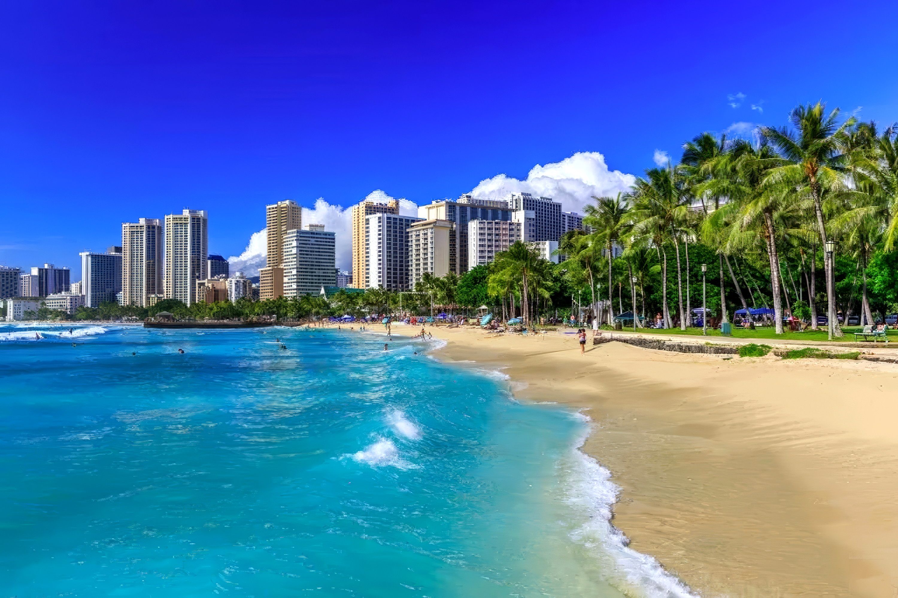 A beach with clear blue water, a sandy shore, and a city skyline in the background.