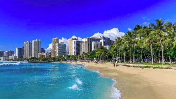 Ein Strand mit klarem, blauem Wasser, Palmen und einer Stadtansicht im Hintergrund.