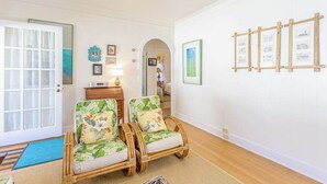 Una habitación con una puerta, dos sillones de mimbre con cojines estampados, una mesita con un radio y cuadros en la pared.