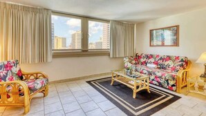 Uma sala de estar com um sofá, duas poltronas, uma mesa de centro e vista para edifícios altos através de grandes janelas.