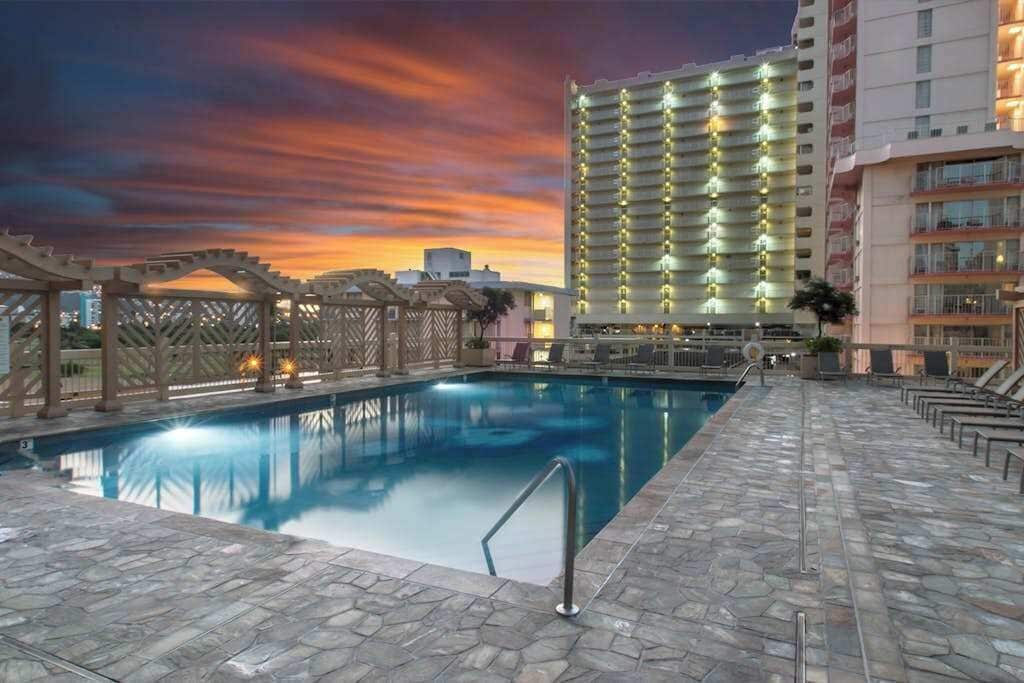 A hotel pool area with a clear view of the pool, a pergola, and a high-rise building in the background.