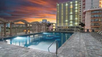 A hotel pool area with a clear view of the pool, a pergola, and a high-rise building in the background.