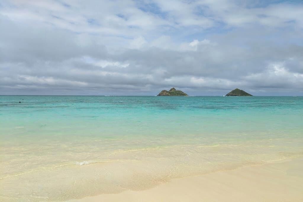A beach with clear turquoise water and distant islands under a cloudy sky.