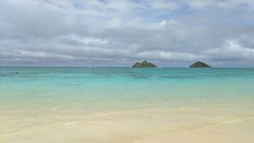 A beach with clear turquoise water and distant islands under a cloudy sky.