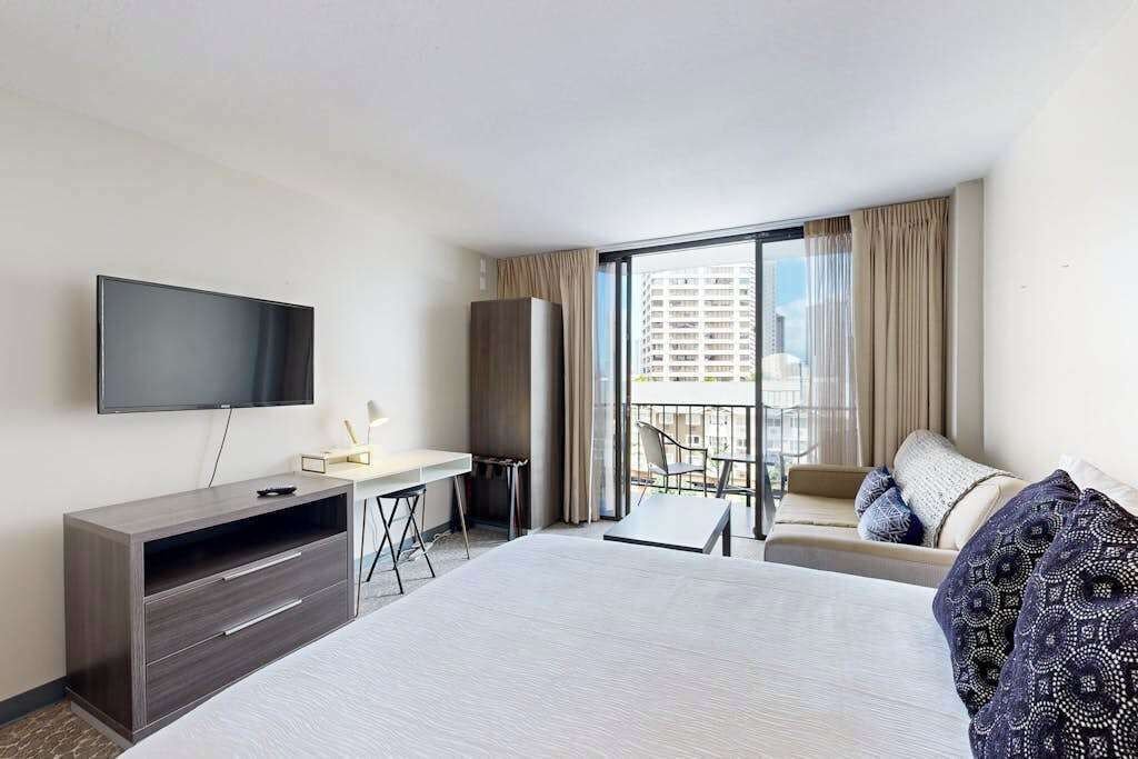 Ein Hotelzimmer mit Bett, Schreibtisch, Fernseher und einem Balkon mit einer Sitzecke, mit Blick auf Gebäude.