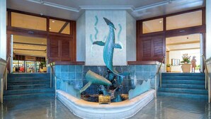 Um lobby com uma escultura de fonte central, uma escadaria e vista para uma loja ao fundo.