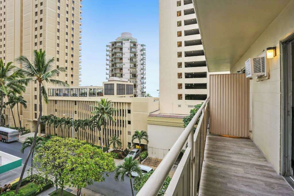 A balcony with a view of tall buildings, a palm tree, and a bus in the background.