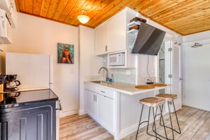 Eine kompakte Küche mit weißen Schränken, einer Mikrowelle und einer Kaffeemaschine. Es gibt Hocker an der Arbeitsplatte und eine Holzbalkendecke.
