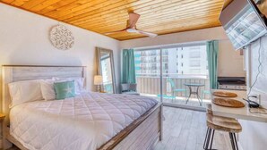 Ein Schlafzimmer mit Bett, Deckenventilator, großem Fenster mit Vorhängen, einem Balkon und einer Küchenzeile mit Herd.