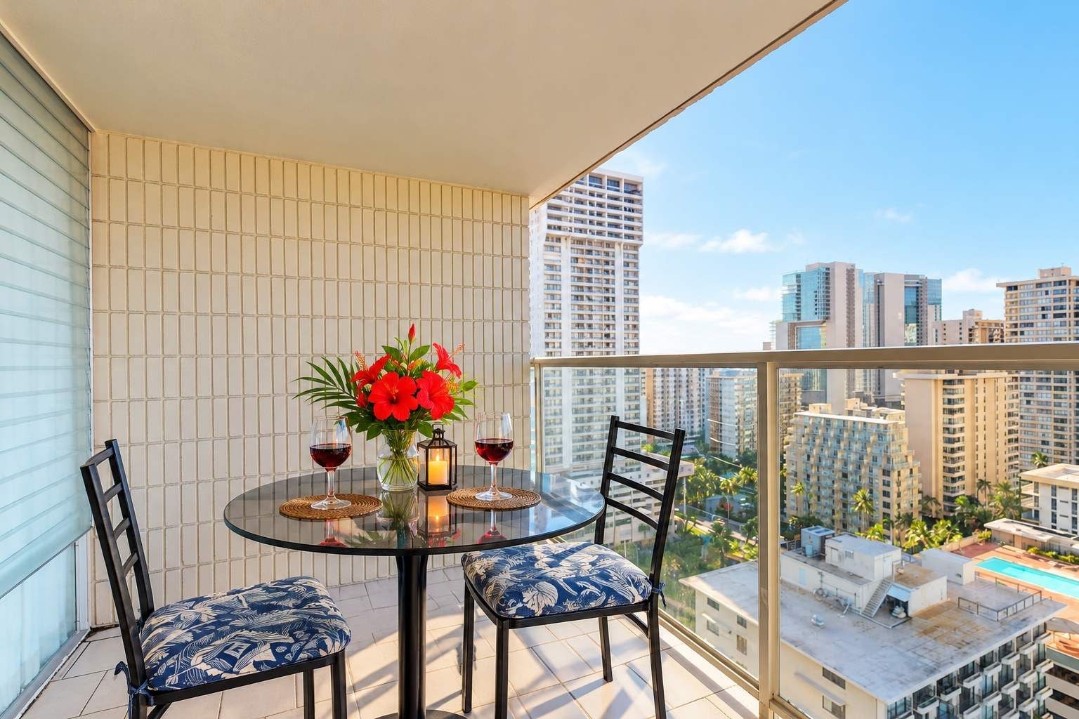 A balcony with a round table, two chairs, a vase with flowers, two glasses of red wine, and a view of a cityscape with tall buildings.