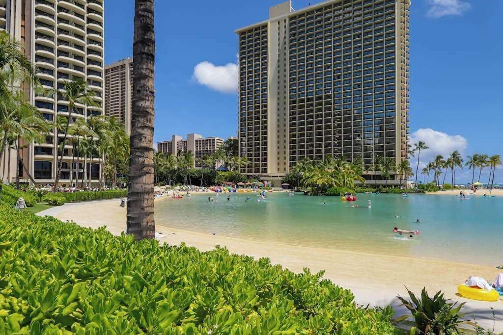 Un hotel frente a la playa con piscina, palmeras y edificios altos al fondo.