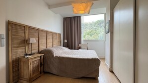 Quarto com cama, mesa de cabeceira, luminária e vista para o verde pela janela.