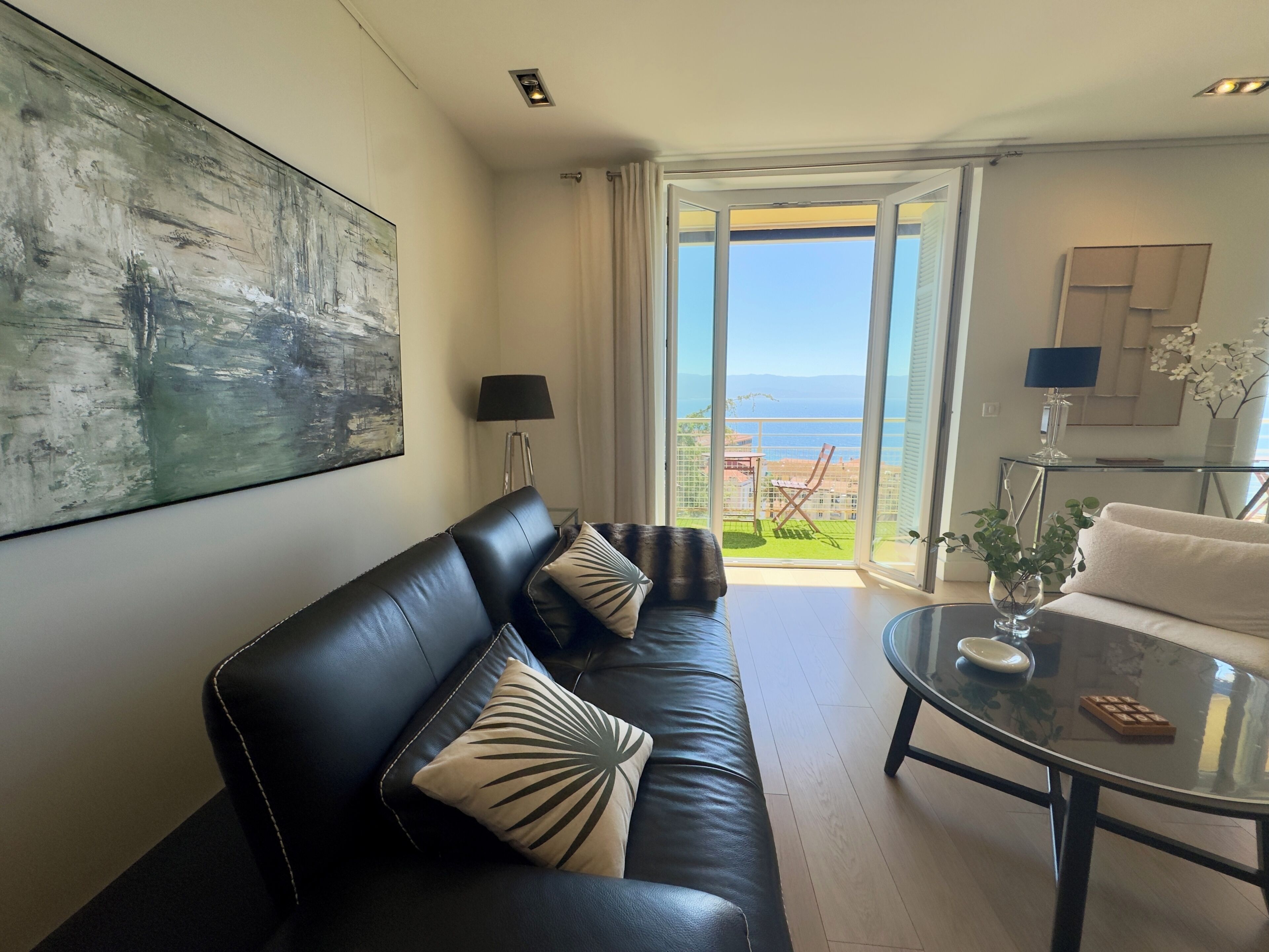 Un salon moderne avec un canapé en cuir noir, une table basse en verre et une vue sur la mer grâce à de grandes portes vitrées.