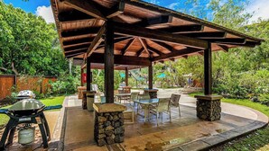 Un patio exterior con una pérgola de madera, pilares de piedra y una parrilla.
