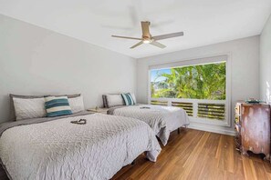 Un dormitorio con dos camas, un ventilador de techo, una ventana con vistas a la vegetación y un aparador de madera.