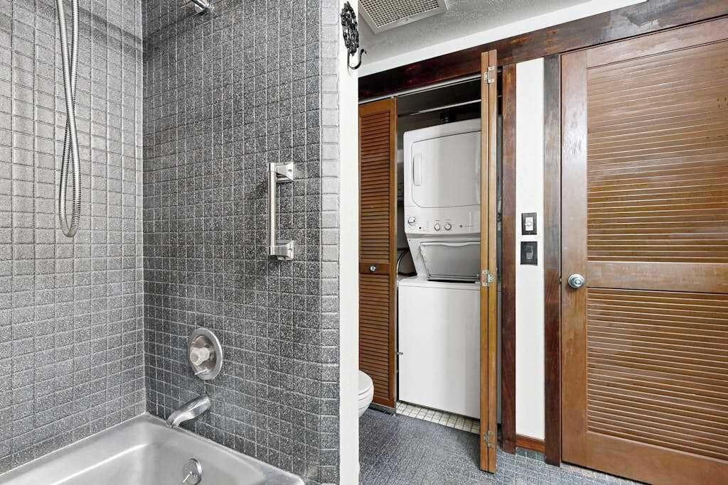 Ein Badezimmer mit Dusche, Waschbecken und Toilette. Eine Holztür führt zu einem Waschraum mit einer Waschmaschine.