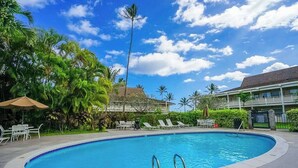 Una piscina con sillas de descanso y sombrillas, rodeada de palmeras y un cielo despejado.