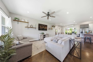 A spacious living room with a large sofa, a coffee table, and a ceiling fan. There is a kitchen area with a refrigerator and a dining table. - The Palm House Family Retreat at Princeville - Mins to Beach, Shops, Dining (Princeville)