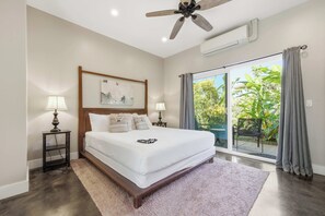 A bedroom with a bed, two bedside tables with lamps, a ceiling fan, a large window with curtains, and a painting on the wall. - The Palm House Family Retreat at Princeville - Mins to Beach, Shops, Dining (Princeville)