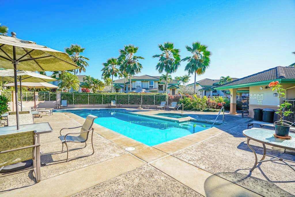 A swimming pool with lounge chairs and umbrellas, surrounded by palm trees and a fence.
