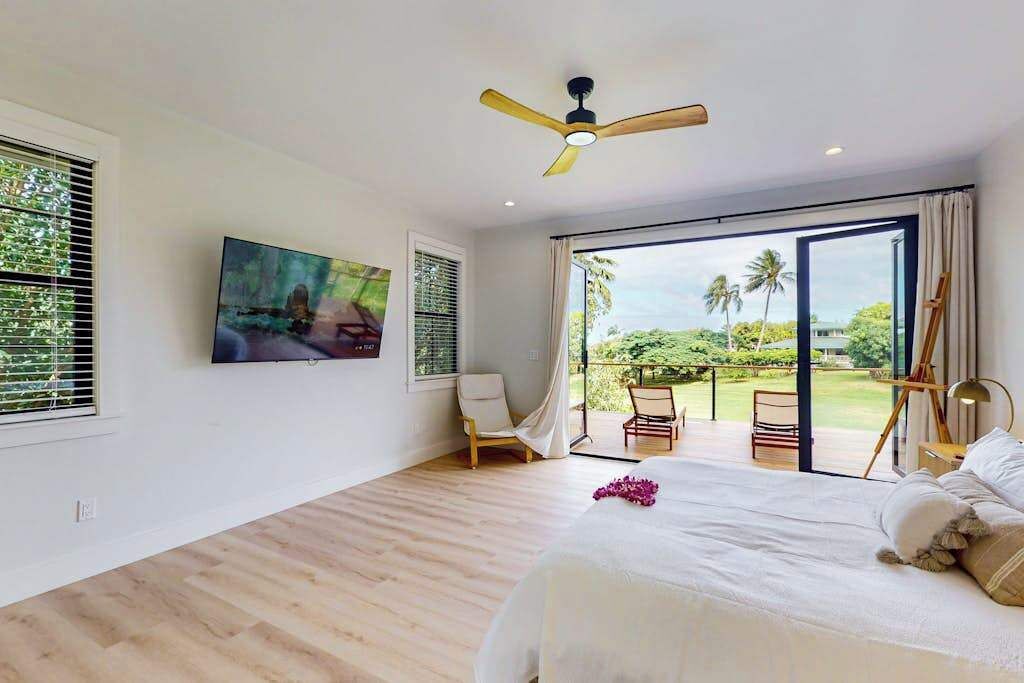 Ein Schlafzimmer mit einem großen Bett, einem Deckenventilator, einem Gemälde, einem Sessel und einem Balkon mit Blick.