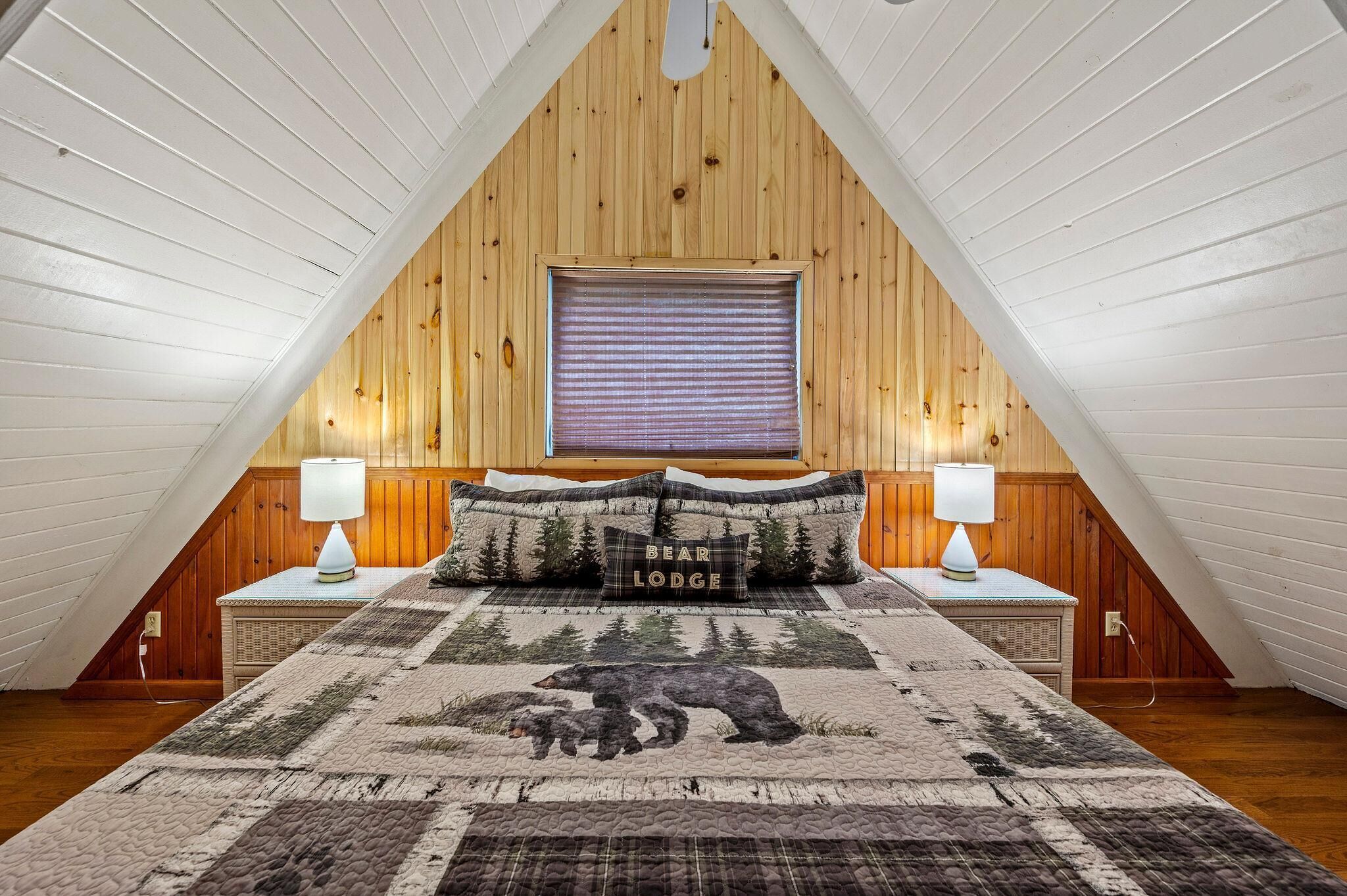 A bedroom with a sloped ceiling, a bed with a quilt, two bedside lamps, a window with blinds, and a pillow with 'BEAR LODGE' text.