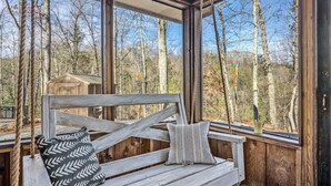 A wooden swing bench with two pillows, large windows, and a view of trees. - Grassy Paradise (Gatlinburg)
