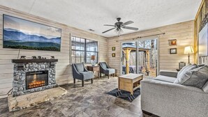 A cozy living room with a stone fireplace, comfortable seating, a ceiling fan, and a large window. - Grassy Paradise (Gatlinburg)