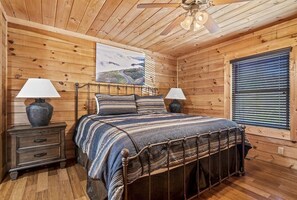 A wooden cabin bedroom with a bed, two bedside tables, a ceiling fan, and a window with blinds. - Happy Days (Gatlinburg)