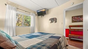 Un dormitorio con cama, televisor, ventana con cortinas, un adorno de oso montado en la pared y un banco rojo.