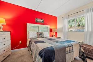 Ein Schlafzimmer mit roter Wand, einem Bett mit Patchworkdecke, einem Nachttisch, einer Lampe, einer Kommode und einem Fenster mit Vorhängen.