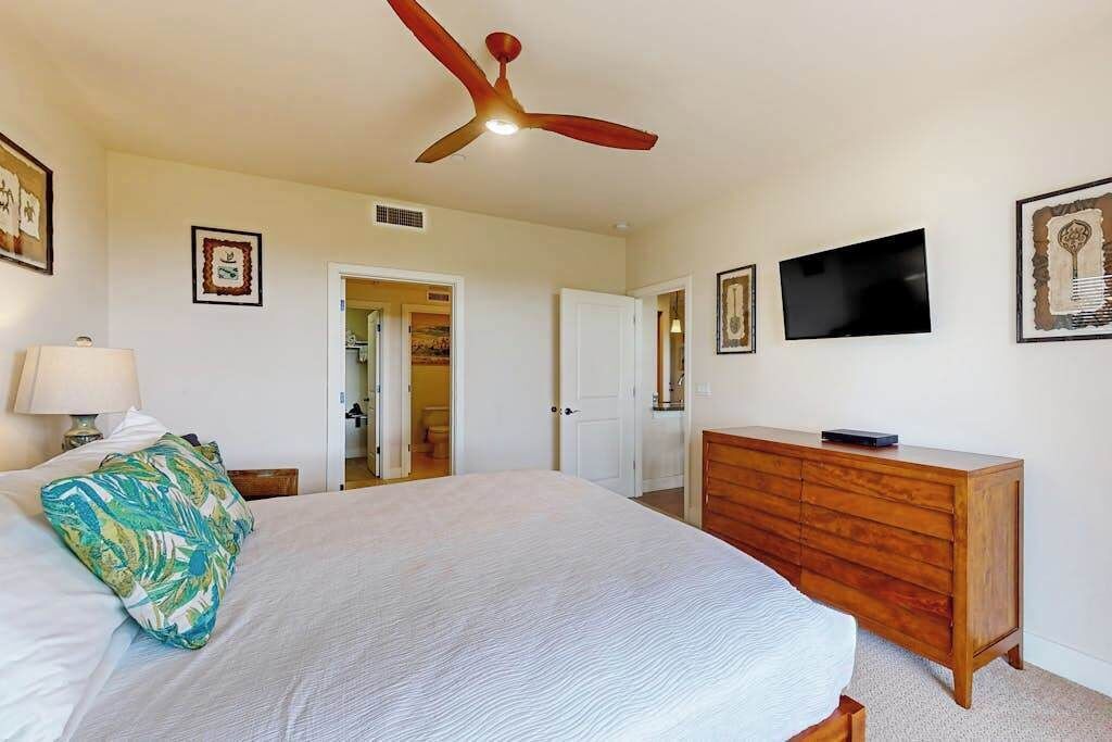 A bedroom with a bed, a wooden dresser, a ceiling fan, a television, and framed artwork on the walls.