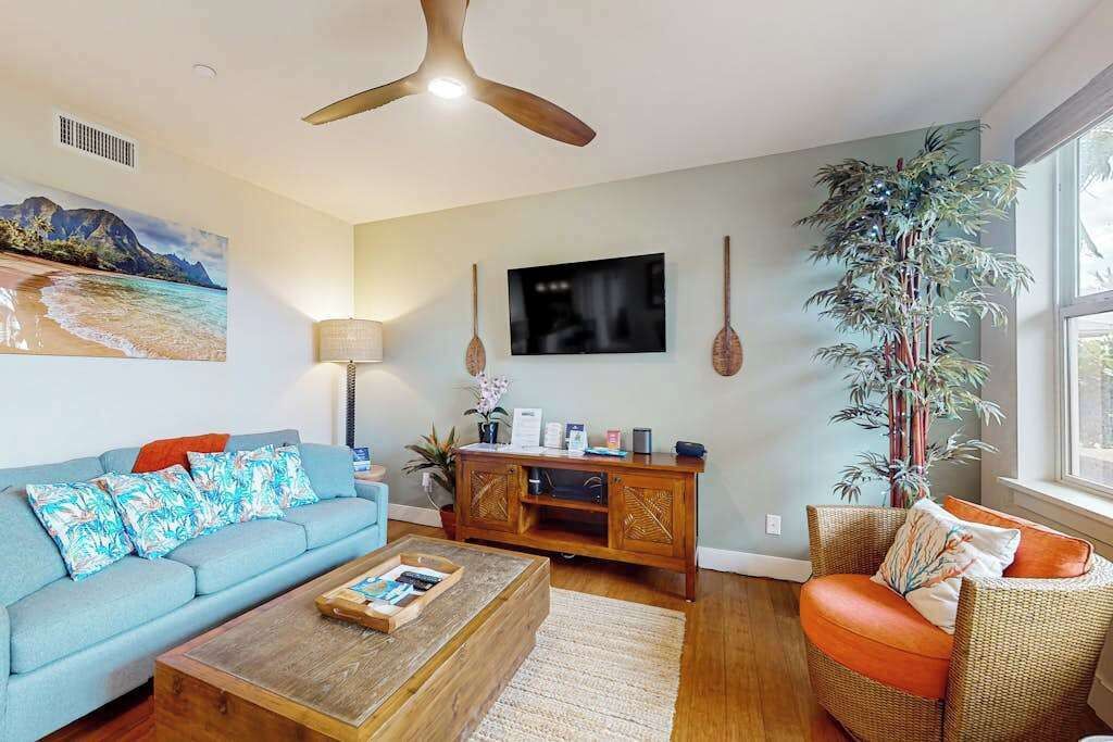 Ein Wohnzimmer mit einer Couch, einem Couchtisch und einem Sessel. Ein Fernseher ist an der Wand befestigt, ein Deckenventilator ist vorhanden und eine große Pflanze steht dort.
