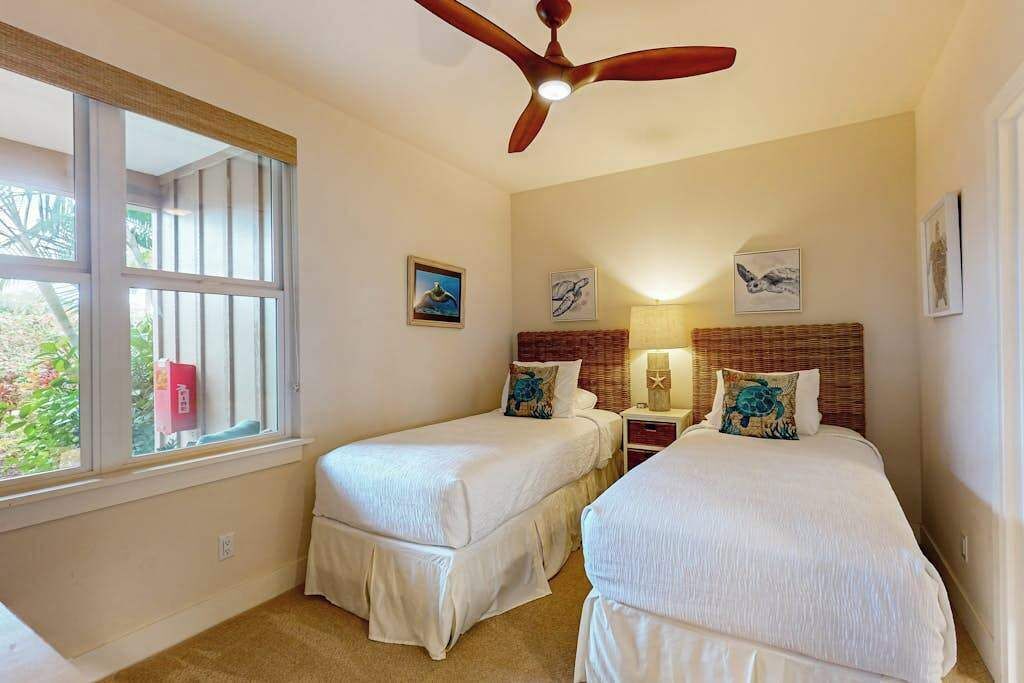 Une chambre avec deux lits, un ventilateur de plafond, une fenêtre avec des barreaux et des œuvres d’art accrochées aux murs.