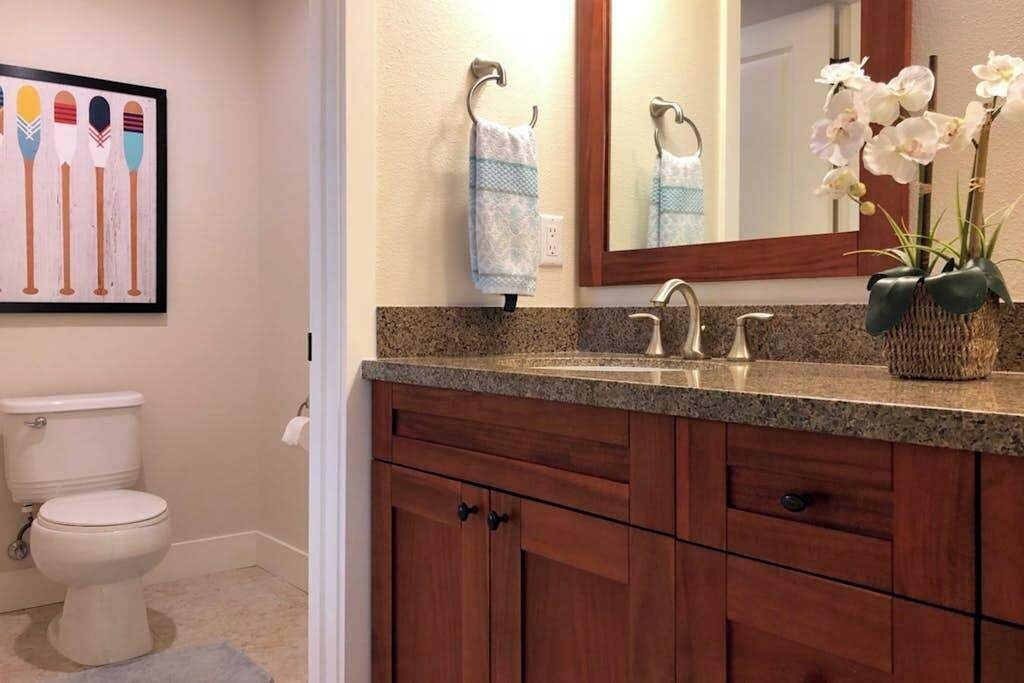 Un baño con encimera de granito, inodoro blanco, armarios de madera, un espejo y una obra de arte enmarcada.