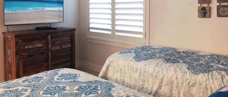 A bedroom with a TV, a window with blinds, and a bed with patterned bedding.