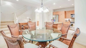 Uma sala de jantar com mesa de vidro cercada por cadeiras de vime, um lustre e uma cozinha ao fundo.