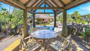 Uma área de estar coberta ao ar livre, com mesa e cadeiras, piscina e paisagismo tropical.