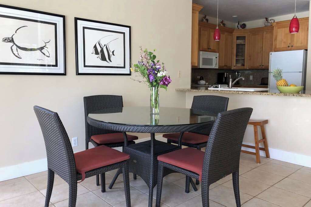 A dining area with a round table, four chairs, a vase with flowers, framed artwork on the wall, and a kitchen in the background.