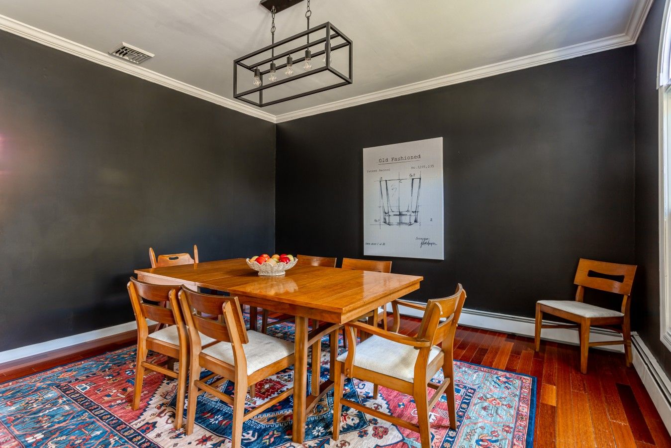 Une salle à manger avec une table et des chaises en bois, un tapis à motifs et un mur noir orné d’une affiche.