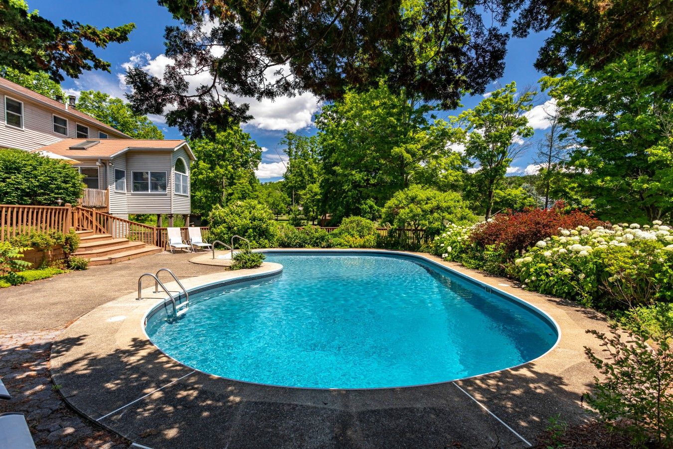 Une piscine à bords incurvés, entourée d’une végétation luxuriante et d’une maison dotée d’une terrasse.