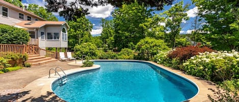 A swimming pool with a curved edge, surrounded by lush greenery and a house with a deck.
