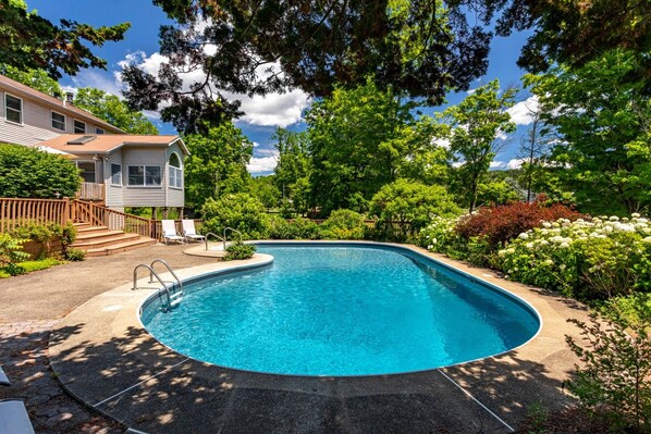 A swimming pool with a curved edge, surrounded by lush greenery and a house with a deck.