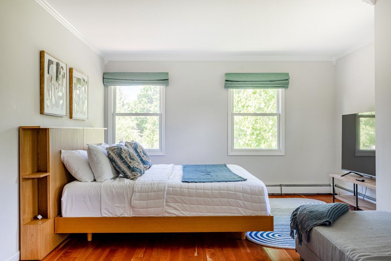 A bedroom with a wooden bed, two windows with green valances, a television on a stand, and a small wooden shelf.