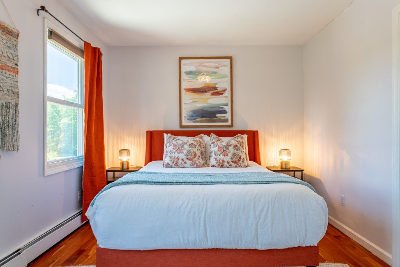Une chambre à coucher avec un lit, deux lampes de chevet, une fenêtre avec des rideaux, un tableau au mur et un radiateur.