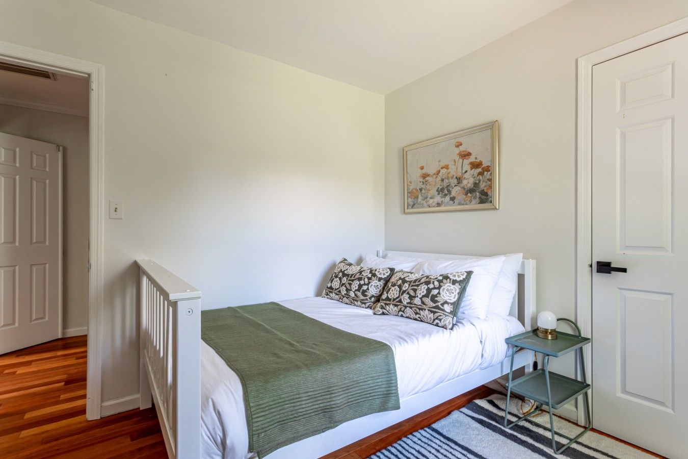 A bedroom with a bed, a green throw blanket, a small bedside table with a lamp, a framed floral artwork on the wall, and a door to the left.
